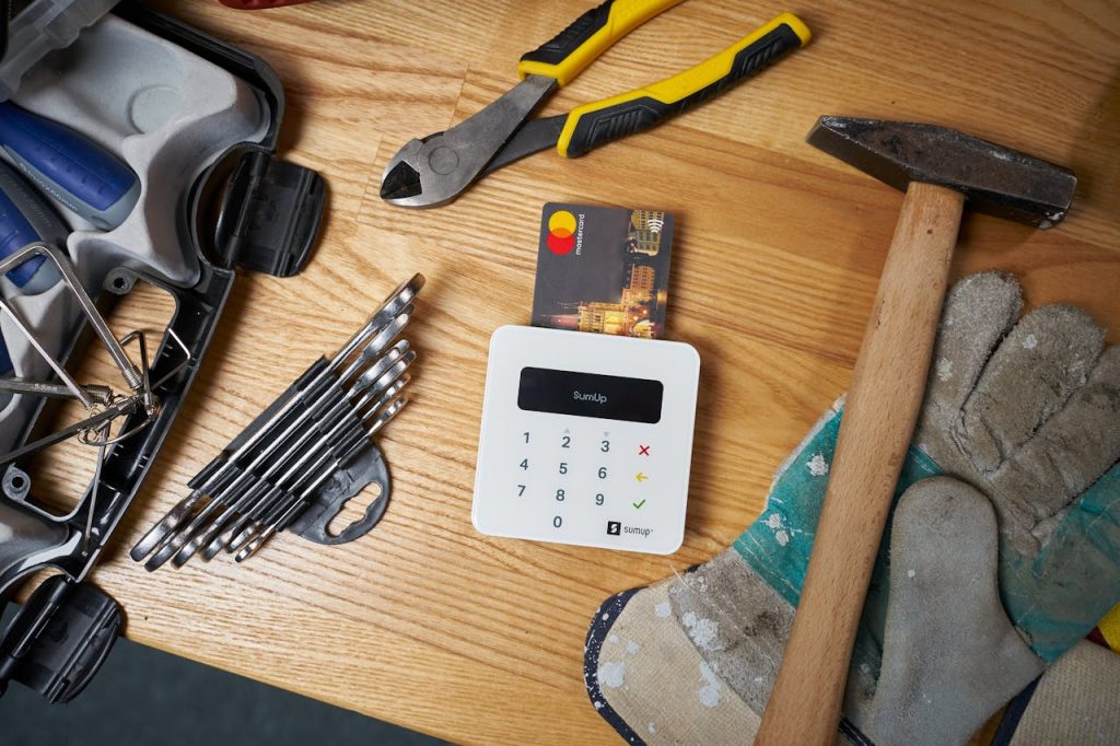 Overhead view of bright small plastic card in white contemporary wireless cash register surrounded by screwdriver set and hammer on big dirty gloves next to pliers